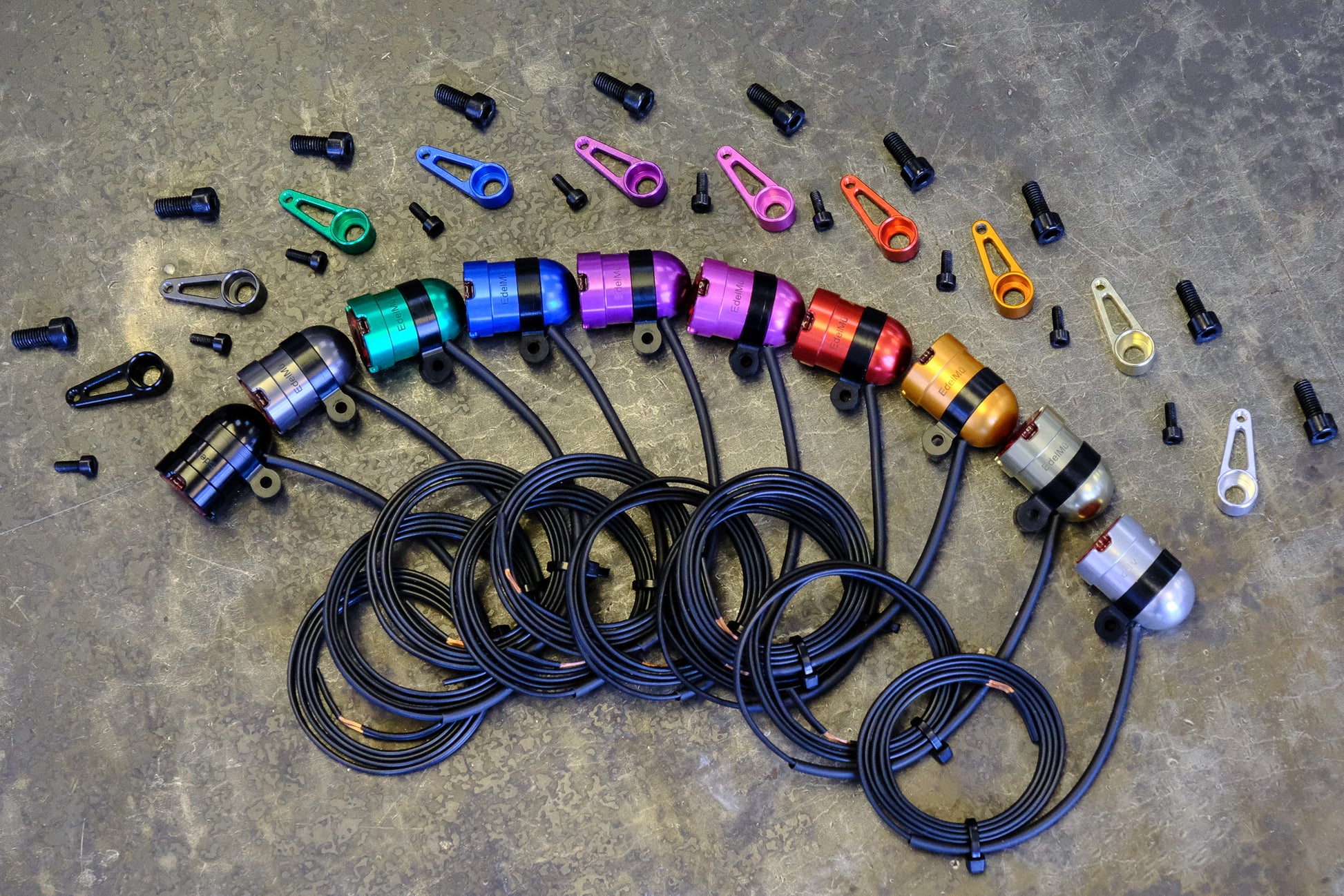 A collection of colorful ALLYGN EdelMü Ring Mounts arranged in a row with matching mounting bolts on a textured gray surface, showcasing lightweight bike accessory components.